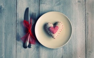 A dinner set-up: A white plate with a heart-shaped pillow on it, a fork and knife tied up with a red ribbon, against a wooden table