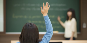 A student in a classroom raises their hand while their teacher stands at the chalkboard