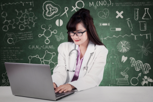 Doctor sits at a laptop in front of a chalkboard with scientific writing on it