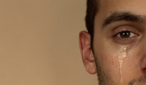 A person is standing in front of a maple colored background with only half of their face showing on the right side of the image. They have a tear falling down their cheek.