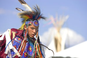 A dancer wearing multicolored beaded clothing and headdress is looking down in mid-dance.