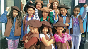 A group of 9 girls wearing brown berets, raising their fists or crossing their arms
