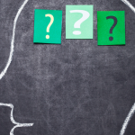 A human head is drawn on a chalkboard. In the brain are three slips of green paper with question marks on them.