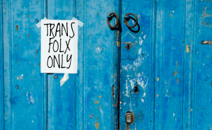 A piece of paper is taped to an antique blue door, which reads "Trans Folx Only."