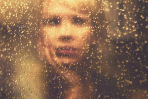 A person is looking at their reflection in a rainy window, touching their face