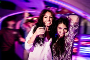 Two people smiling and standing near each other in a club, one holding a drink and the other with their arm raised behind their head.