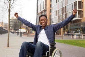 A person sits in a wheelchair, stretching out their arms and smiling.