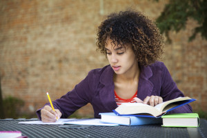 A person is sitting outdoors, writing on a pad of paper as their hand rests on a stack of books.