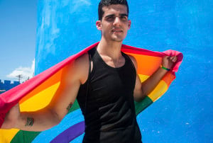 A person stands confidently staring into the camera with a rainbow flag draped over their shoulders.
