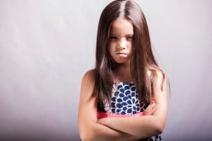 A child with long brown hair stands with their arms folded, pouting.