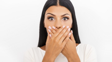 Shocking news. Surprised young woman covering mouth with hands and staring at camera while standing against white background