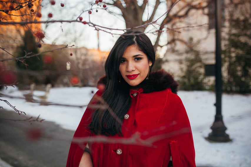 The photo features a woman with long hair and a bright red coat standing in a snowy park. The woman in the photograph, Patricia Valoy, is a Latina feminist.