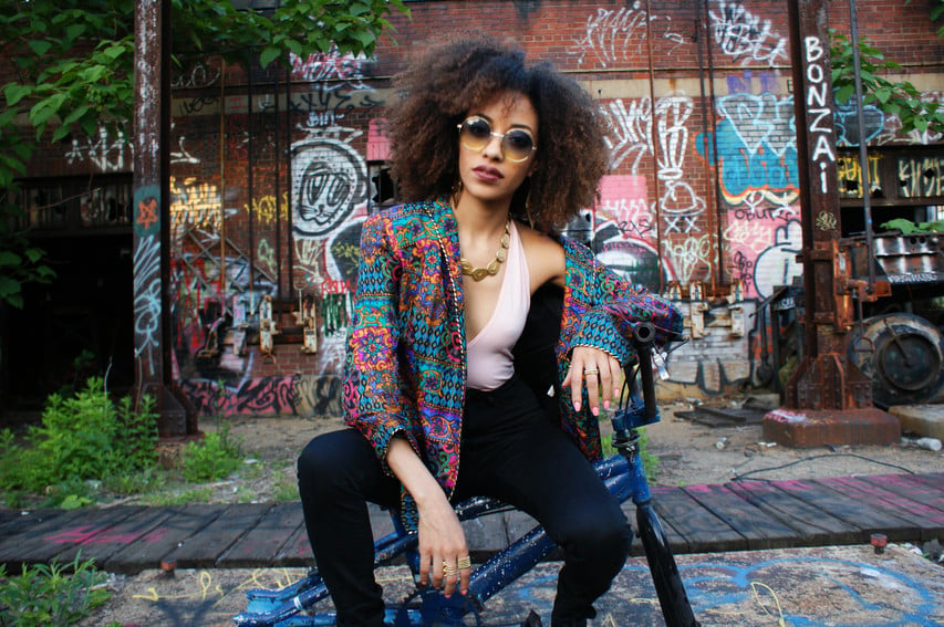 The image features a black Latina woman, sitting on a bicycle with a colorful jacket and round sunglasses. She has a shoulder-length afro.