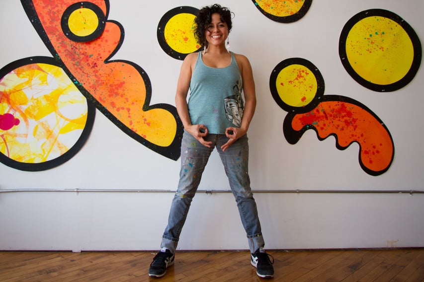 The picture features a Latina woman with short hair standing in front of a bright piece of artwork – different shapes of reds, oranges, and yellows.