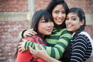 Three friends are hugging one another outside, standing against a red brick wall.