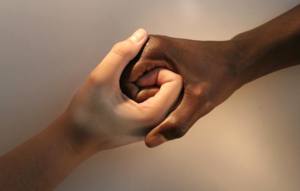 Two hands – one with a light skin tone, one with a dark skin tone – are holding each other.