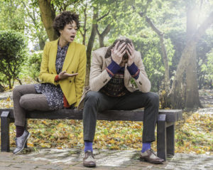 Two people sit on a bench in the park, arguing. One of them is gesturing to the other, who has their face covered with their hands.