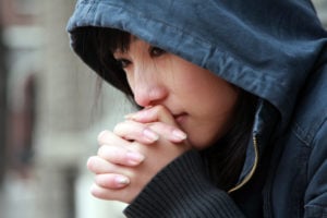 A person wearing a dark-hooded jacket appears worried, with their hands laced and touching their face.