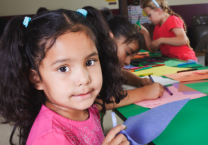 A child, doing crafts with construction paper, gazes into the camera with a half-smile. Two other children are working behind them.