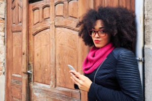 A person with an afro gazes into the camera while wearing red glasses, a pink infinity scarf, and a black jacket.