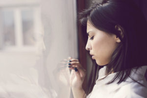 A person with long black hair stands in front of a window, their hand lightly touching it with a pained expression.