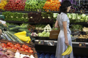 A person stands in a grocery store, in an aisle filled with colorful vegetables, holding a purse and shopping basket in one hand and a bunch of bananas in the other.