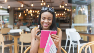 A person sitting outdoors at a cafe holds up a bright pink tablet to see their reflection as they hold up a tube of lipstick near their face.