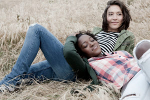 Two young people lying in a grassy field – one with their head resting on the stomach of the other.