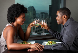A couple sits across from one another, toasting with wine glasses, with two plates of food on the table before them.