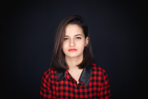 A person staring straight ahead with a red plaid shirt and bright red lipstick against a black background.