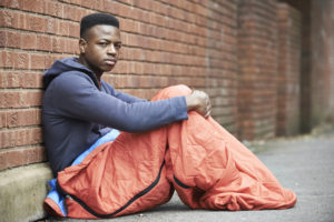 A young person sits on the ground against a red brick wall, half-inside a bright orange sleeping bag.