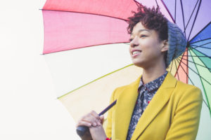 A person smiling as they stand beneath a rainbow umbrella.