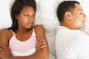 Two people in bed together. One is on their side with their eyes closed; the other person has their arms folded, staring at the person next to them with dissatisfaction.