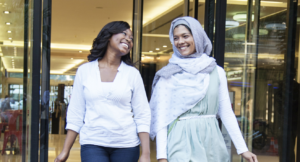 Two people – one wearing a head scarf and one without – walk out of a shopping mall, smiling at each other.