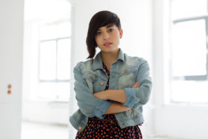 Young person with an asymmetrical haircut and a denim jacket stands in front of two windows with their arms crossed.