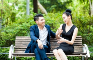 Two people sitting on a bench. One turns to face the other, smiling and listening intently. The other is talking with a skeptical face, moving their hands to gesture something.