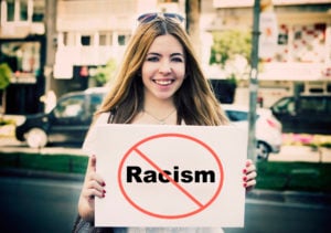 A person standing in a street, smiling and holding a white placard with the word "Racism" in a cross-out circle. 