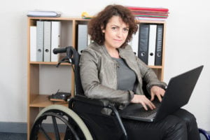 A person sitting in a wheelchair with a laptop on their lap, grinning slightly at the camera.