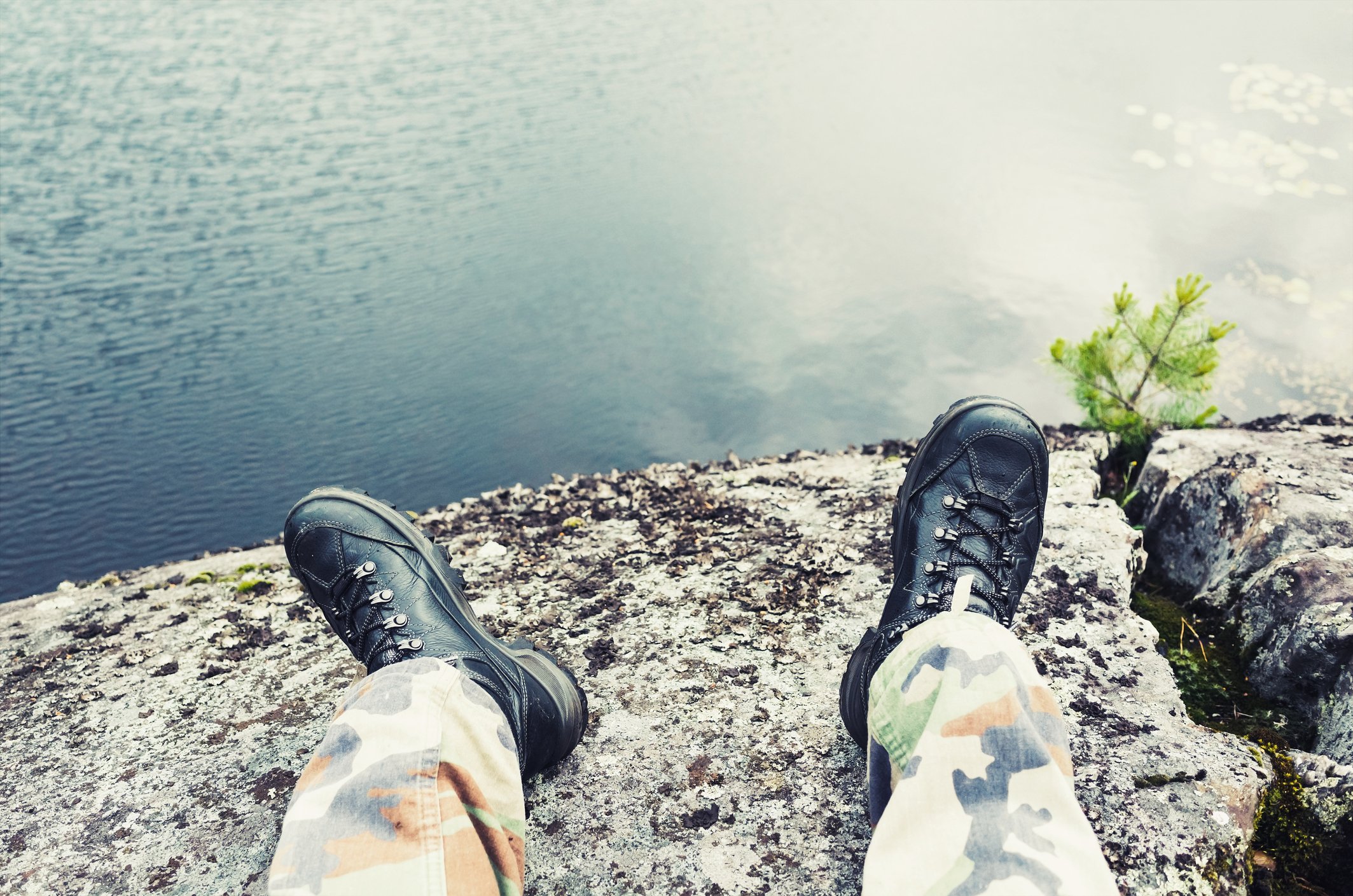 Two legs in camouflage pants and black boots, sitting near a body of water.