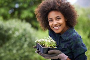 A person holding a tray of seedlings in a garden, smiling.