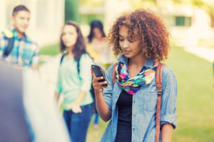 A teen looks down at their cell phone while standing outdoors, with other people out of focus behind them.