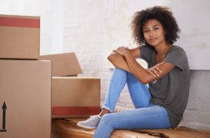 A person sits with a half-smile, one knee drawn to their chest, by a stack of boxes.