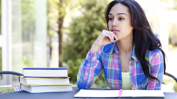 A student sits at an outdoor table with books stacked beside them, looking contemplative.