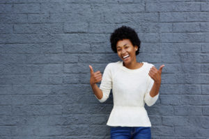 A young person standing against a grey brick wall, gesturing a "thumbs up" sign with each hand.