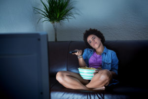 A person sits alone with popcorn, tilting their head and pointing a remote control at a television.
