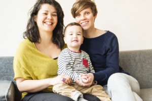 Family portrait of a queer couple sitting on a couch together with a small child on their laps.