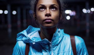 Closeup of a person who appears worried, their glance turned upward, as they walk a city street at night with headphones in their ears.