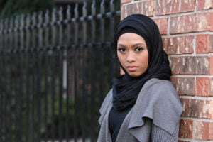A person wearing hijab leans against a brick wall.