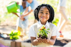 A young student holds up a pepper plant while smiling into the camera. Students in the background are out of focus, using gardening tools.