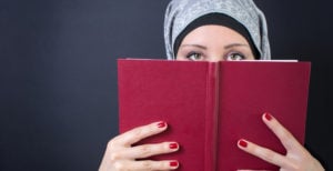 A person wearing hijab, holding a book to cover their face.